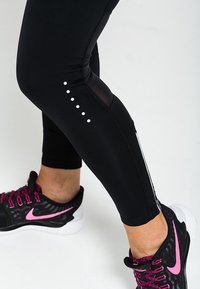 Black leggings featuring a mesh insert and white dot accents along the side. Paired with black and pink athletic shoes.