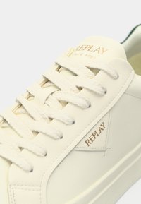 White leather sneaker with textured surface, featuring round laces, a logo in gold on the side, and a padded ankle area for comfort.