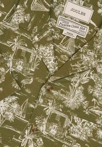 Zelená bavlnená košeľa s bielym tropickým vzorom, ktorý zobrazuje domy, palmy a plachetnice. Obsahuje hnedý gombík a značkovú etiketu.