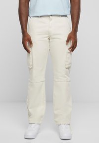 Pantalons cargo beiges avec plusieurs poches latérales, jambes droites et coupe décontractée, associés à des baskets blanches. Fabriqués en tissu en coton durable.