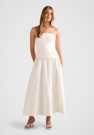 Femme portant un haut blanc sans bretelles et ajusté ainsi qu'une longue jupe blanche fluide, associée à des sandales blanches à talons, debout sur un fond uni.