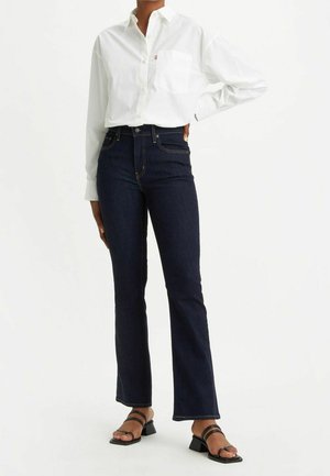 Persona vestida con una camisa blanca impecable de botones, vaqueros de tiro alto azul oscuro y sandalias negras de tacón, de pie sobre un fondo blanco.