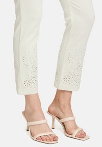 Białe dżinsowe spodnie z haftowanymi kwiatowymi wzorami i kryształkami na krawędzi. Nosi je w połączeniu z cielistymi sandałami na wysokim obcasie ze strunami.