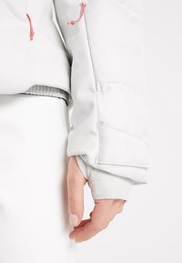 Main portant une manche texturée gris clair s’étendant d’une veste blanche matelassée avec des cordons roses, sur fond blanc.