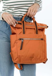 Zaino in nylon arancione con due manici superiori, una tasca con zip anteriore e dettagli in pelle marrone. Texture liscia e design moderno.