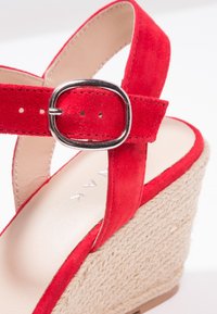 Sandale compensée en daim rouge avec une plateforme en jute tressé. Dotée d'une bride avec boucle argentée et d'une doublure intérieure lisse.