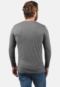 Szürke hosszú ujjú póló kerek nyakkivágással, puha anyagból készült, testhezálló szabású és sima tapintású, kék farmerrel viselve.