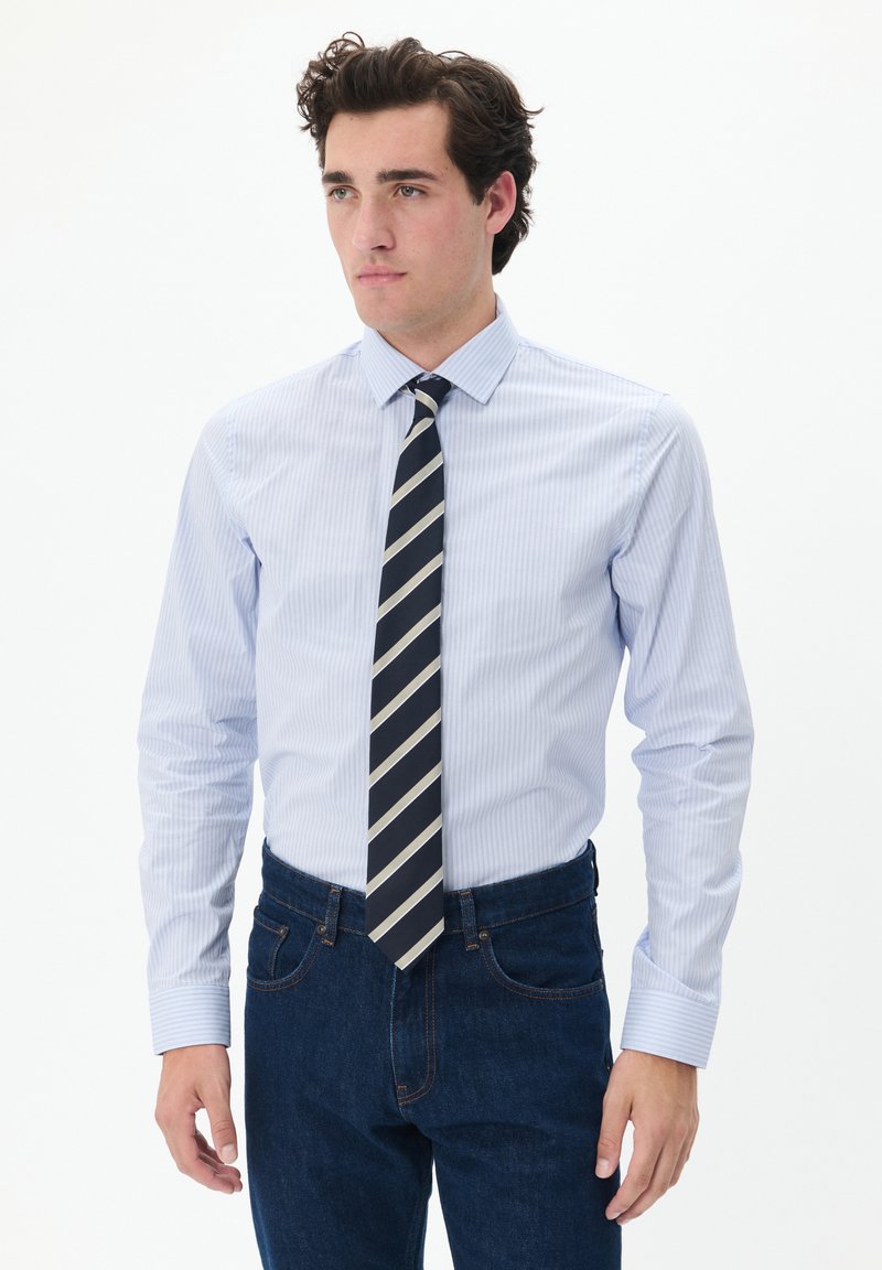 Camisa de rayas azul claro con cuello y mangas largas, acompañada de una corbata azul marino con rayas diagonales en blanco y gris, combinada con vaqueros azul oscuro.