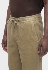 Nahaufnahme einer Person, die beige Cordhose mit elastischem Bund und Kordelzug trägt, kombiniert mit einem braunen Hemd.