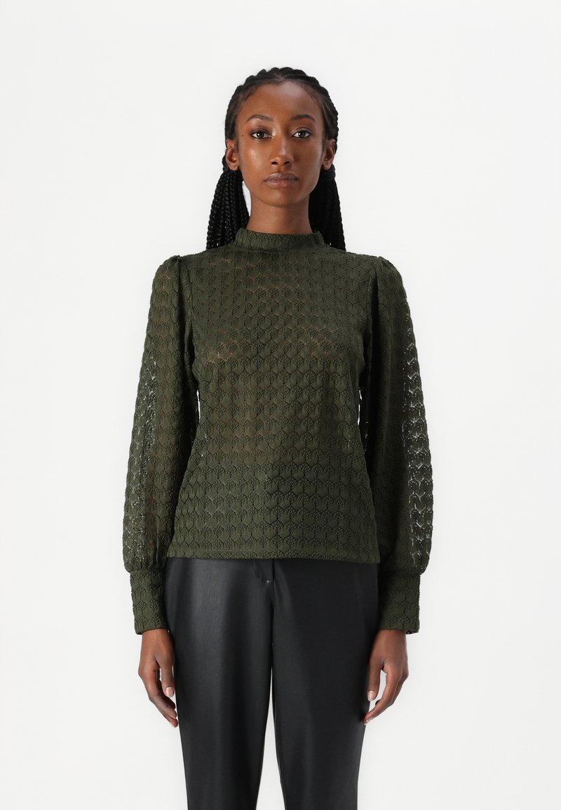 Tamsiai žalia tekstūruota megztinis su ilgomis rankovėmis, aukštu kaklu ir geometriniu raštu, derinamas su juodomis kelnėmis.