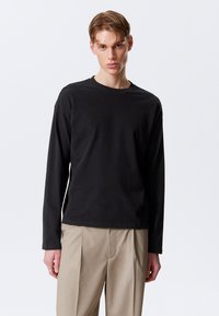 Tricou lung cu mâneci negre, din bumbac, cu guler rotund și o croială lejeră. Materialul pare neted, fără modele sau accente.