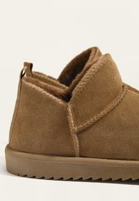 Brązowe zamszowe buty wsuwane z puszystym futerkiem w środku, zaokrąglony czubek i żebrowana gumowa podeszwa, przedstawione z boku na jasnym tle.