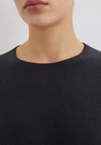 Schwarzer Strickpullover mit rundem Ausschnitt, der eine strukturierte Oberfläche und minimalistische Stickdetails aufweist. Nahaufnahme des Ausschnittbereichs.