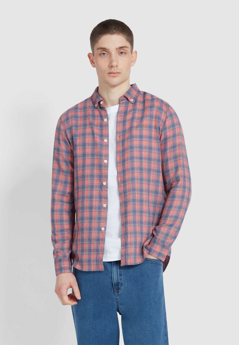 Młody mężczyzna w rozpiętej czerwono-niebieskiej koszuli w kratę na białym T-shircie i niebieskich dżinsach na jasnoszarym tle.