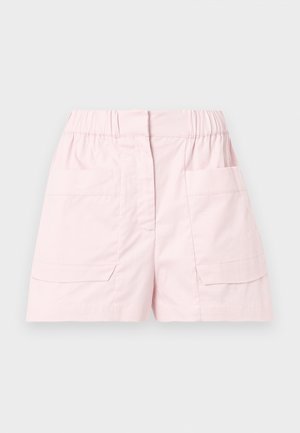 Femme aux longs cheveux foncés debout, les mains sur les hanches, portant un T-shirt blanc orné, un short rose pâle et des mocassins marron foncé à pampilles.