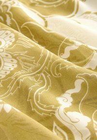 Tissu présentant des motifs floraux blancs délicats et des motifs tourbillonnants sur un fond jaune clair doux, avec une texture subtile et des plis superposés.