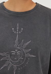 Tmavě šedá mikina s grafikou slunce a měsíce ve světle šedé barvě. Design obsahuje jemné detaily a text s nápisem "BOHEMIA."