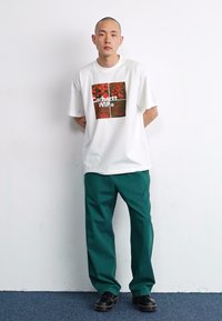White cotton t-shirt featuring a square graphic with red and green patterns; paired with green pants and black shoes.