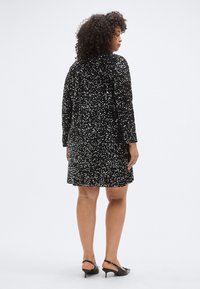 Fekete flitteres ruha hosszú ujjakkal, térd fölötti fazonnal és texturált felülettel. Fekete magassarkú cipővel viselve.