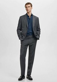 Szary garnitur o dopasowanym kroju, teksturowana tkanina, w połączeniu z ciemnoniebieską koszulą i czarnymi wypolerowanymi butami, ręce w kieszeniach, neutralne tło.