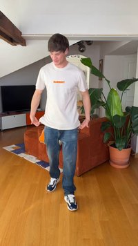 Ung mann kledd i hvit t-skjorte og jeans står i en stue med oransje sofa, stor grønn plante og tregulv.