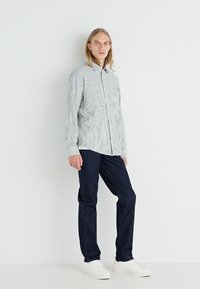 Striped button-up shirt in white and blue, paired with dark denim pants and white shoes. Simple, modern design with a chest pocket detail.