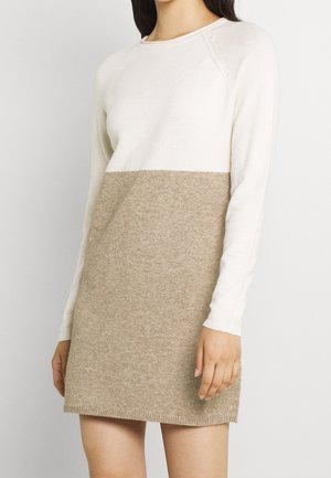 Femme portant une robe tricotée à manches longues bicolore, avec une moitié haute blanche et une moitié basse beige, debout devant un fond uni.