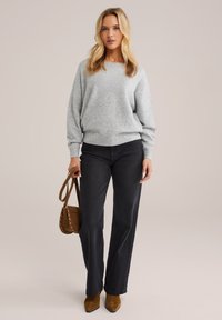 Pulover gri deschis, blugi negri cu picioare largi și botine cu toc din suede maro. Geanta este mică, maro, cu accente cu ținte. Textură moale, design casual.