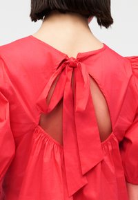 Femme portant une robe rouge à manches bouffantes avec un grand nœud noué dans le haut du dos, dévoilant un détail d'ouverture en forme de trou de serrure.
