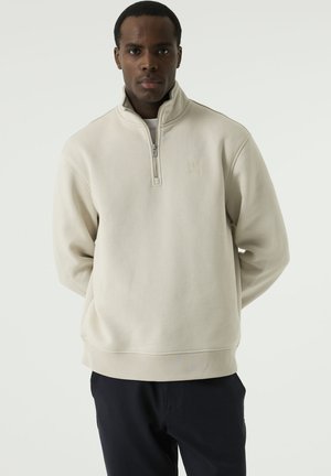 Mann steht mit den Händen hinter dem Rücken, trägt einen hellbeigen Quarter-Zip-Pullover und dunkle Hosen, blickt geradeaus vor schlichtem Hintergrund.