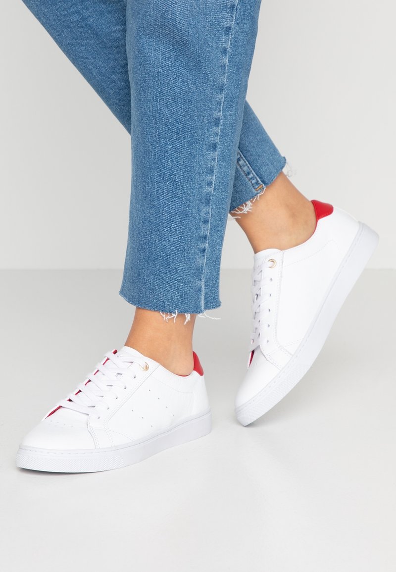 Zapatillas bajas blancas con acentos rojos, parte superior de cuero texturizado, ojetes dorados y detalles perforados. Llevadas con jeans azules arremangados.