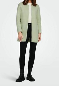 Chaqueta verde claro con textura, de corte recto y mangas largas, combinada con leggings negros y botas negras gruesas.