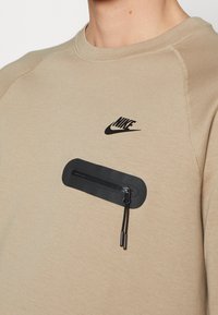 Beżowa bluza Nike z czarnym zamkiem błyskawicznym na piersi i czarnym logo Nike nad kieszenią.