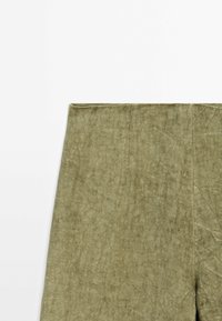 Gros plan sur un tissu texturé vert olive en forme rectangulaire avec des coutures visibles sur un fond blanc.