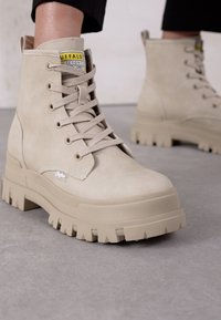 Stivali alla caviglia chunky beige con lacci e suole in gomma spessa, indossati con pantaloni neri su un pavimento di cemento.