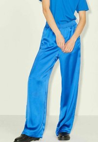 Person wearing shiny blue satin wide-leg pants, blue shirt, and black shoes, standing with hands clasped in front.