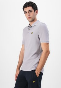 Grå polo skjorte med korte ermer, tre knappestolper og en gul brodert logo på venstre bryst. Bæres med mørk marinebukser.