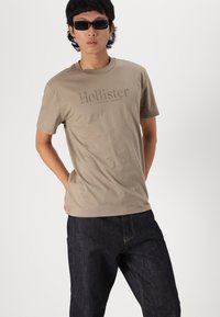 Beige pamut póló sötétebb árnyalatú "Hollister" logóval, rövid ujjakkal és kerek nyakkivágással, sötét farmernadrággal párosítva.