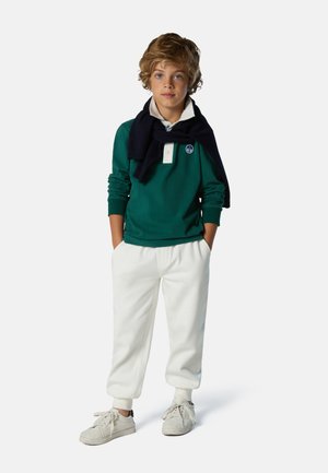 Grünem Langarmhemd mit weißem Kragen und Logo, weißen Sweatpants und beigen Turnschuhen, mit einem marineblauen Pullover um den Hals drapiert.