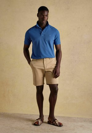 Mann trägt blaues Polohemd, beige Shorts und gemusterte Slip-On-Schuhe, steht vor einer schlichten beigen Wand.