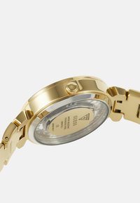 Zlaté metalové hodinky s okrúhlym puzdrom, minerálnym sklíčkom na zadnej strane a nerezovým remienkom. Obsahujú korunku na nastavenie a gravírované detaily.