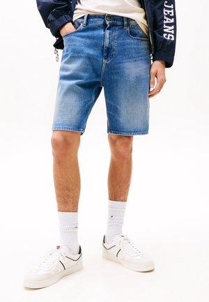 Lichte blauwe denimshorts, knielang, met gestikte zakken, een knoopsluiting en een subtiele vervaagde textuur. Gecombineerd met witte sneakers en sokken.