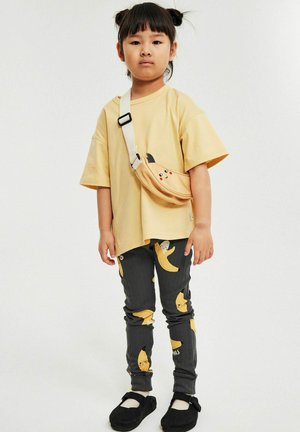 Kind mit schwarzen Haaren in Dutts trägt ein gelbes übergroßes Hemd, graue Hosen mit Bananenmuster, schwarze Schuhe und eine gelbe Umhängetasche mit einem Gesicht.