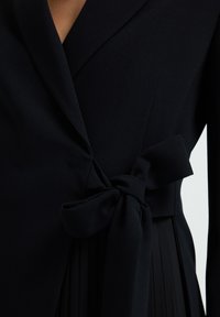 Schwarze taillierte Jacke mit tiefem V-Ausschnitt und großer Stoffschleife, die um die Taille gebunden ist, über einem plissierten Rock oder Kleid.