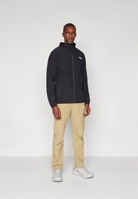 Juoda lengva striukė su užtrauktuku priekyje, kapišonu ir šoninėmis kišenėmis, derinama su smėlio spalvos lauko kelnėmis ir pilkais sportbačiais.