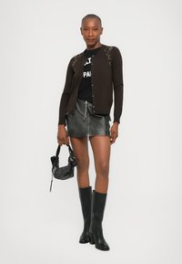 Bruine cardigan met knoopsluiting en bloemborduurwerk, gedragen over een zwarte graphic tee, gecombineerd met een grijze leren minirok en zwarte knielange laarzen.