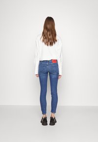 Plave skinny traperice s narančastom oznakom koja sadrži "HUGO", u kombinaciji s bijelom košuljom dugih rukava i crnim cipelama.