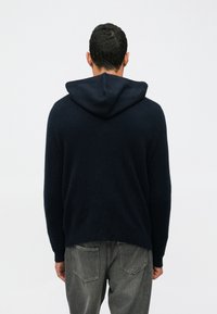 Vyras, apsirengęs tamsiai mėlynu kapriu ir išblukusiomis juodomis džinsomis, stovi pasukęs nugarą į šviesų baltą foną.