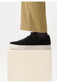 Pied portant une basket basse noire avec une semelle blanche et un pantalon beige, posé sur un bloc beige sur fond blanc.