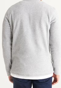 Grå fleece-sweatshirt med rund halsudskæring, struktureret overflade og ribbede manchetter, båret over en hvid lagdelt skjorte, kombineret med denim.